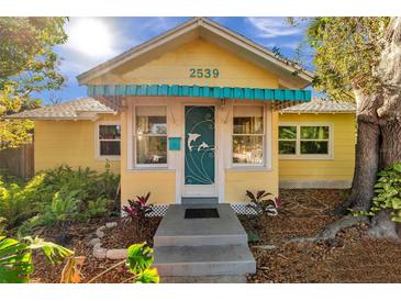 Charming yellow bungalow featuring a whimsical door, bright blue awning, and professionally landscaped front yard at 2539 44Th S St, St Petersburg, FL 33711