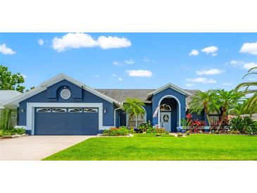 Charming blue home featuring a two-car garage, lush green lawn, and colorful landscaping under a bright, partly cloudy sky at 28390 Openfield Loop, Wesley Chapel, FL 33543