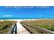 Inviting beach view featuring a wooden walkway to the sandy shore, surrounded by lush vegetation and clear blue skies at 4133 Heron Way # E202, Bradenton, FL 34205