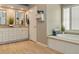 Bathroom with dual sinks, white cabinetry, a soaking tub, plantation shutters, and wood-look flooring at 5949 225Th E St, Bradenton, FL 34211