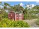 View of a red storage shed, with white trim, surrounded by tropical foliage and located on a gravel area at 730 Buckskin Ct, Englewood, FL 34223