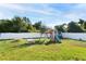 Backyard with green grass, white fence, playset, and mature trees under a bright blue sky at 2408 Bonanza Ln, North Port, FL 34286