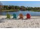 Four Adirondack chairs sit on a sandy beach, facing a serene lake surrounded by lush greenery at 4123 Cascade Falls Dr, Sarasota, FL 34243