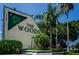 Entrance sign for 'The Woods' community featuring lush greenery and palm trees under a clear blue sky at 39650 Us Highway 19 N # 1422, Tarpon Springs, FL 34689