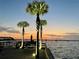 Waterfront view from the pier features lit palm trees, a beautiful sunset and sailboats enhancing the property's appeal at 100 Bluff View Dr # 117C, Belleair Bluffs, FL 33770