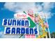 Sign for Sunken Gardens featuring blue, pink, white and green colors with various tropical plants framing the sign at 12924 Gulf Blvd # 310, Madeira Beach, FL 33708