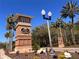 Community entrance featuring a decorative welcome sign, brick accents, and lush landscaping with palm trees at 5051 Gevalia Dr, Brooksville, FL 34604