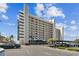A bright exterior shot of a tall condominium with a parking lot and palm trees, set against a partly cloudy blue sky at 15462 Gulf Blvd # 504, Madeira Beach, FL 33708