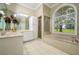 Bright bathroom featuring double sinks, white cabinetry, a walk-in shower, and a large soaking tub under an arched window at 8444 Shenandoah Run, Wesley Chapel, FL 33544