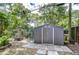A gray backyard shed with double doors sits on a stone patio surrounded by mature landscaping and a colorful fence at 2051 Indigo Dr, Clearwater, FL 33763