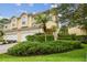 A two-story yellow townhome featuring a two-car garage and lush landscaping with palm trees and flowering bushes at 3484 Primrose Way, Palm Harbor, FL 34683