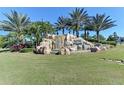 Community entrance featuring a waterfall and lush landscaping with palm trees and manicured lawn under a bright blue sky at 115 Winding River Trl, Bradenton, FL 34212