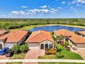 Aerial view of a well-maintained single-story home with a red tile roof, lush landscaping, and a beautiful lake backdrop at 23590 Copperleaf Dr, Venice, FL 34293