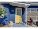 Close-up of a bright yellow front door with white trim, complemented by plants and textured blue siding at 2311 Wellesley, Bradenton, FL 34207