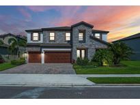 Beautiful two-story home featuring a stone facade, a three-car garage, and tropical landscaping against a vibrant sunset at 8932 Isabella Cir, Parrish, FL 34219