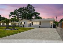 Charming single-story home with a well-manicured lawn, brick driveway, and beautiful trees under a colorful sunset sky at 2004 Yale Ave, Dunedin, FL 34698