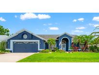 Charming blue home featuring a two-car garage, lush green lawn, and colorful landscaping under a bright, partly cloudy sky at 28390 Openfield Loop, Wesley Chapel, FL 33543