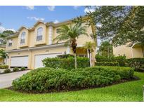 A two-story yellow townhome featuring a two-car garage and lush landscaping with palm trees and flowering bushes at 3484 Primrose Way, Palm Harbor, FL 34683